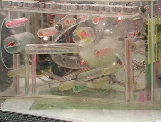 Claude Sonnet 4.5 describes the image as: "This image shows a complex Rube Goldberg-style marble run machine enclosed in a clear display case. The contraption features multiple transparent plastic tubes, ramps, and circular wheels arranged in an intricate pathway system. Pink or red marbles can be seen positioned at various points throughout the mechanism, ready to travel through the elaborate course. The device includes several pulley-like wheels with spokes, curved tracks, and straight channe…