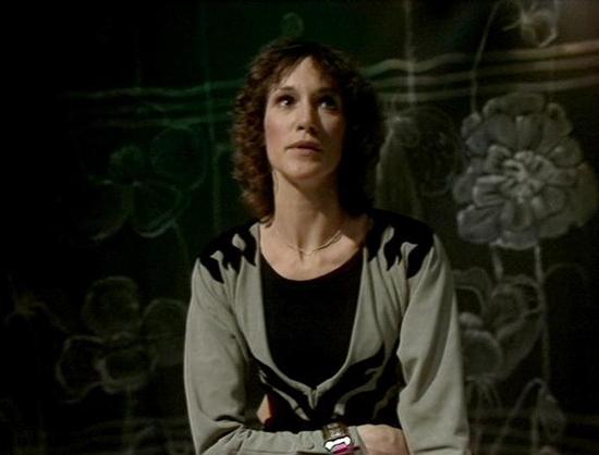 GPT 4.1 Mini describes the image as: "The image shows a woman with short, curly brown hair, wearing a light gray cardigan with black patterns over a black top. She is seated or standing against a dark background featuring some artistic, indistinct floral or abstract designs, giving the scene a somewhat introspective or somber mood. The lighting is soft but focused on her face, highlighting her expression, which appears thoughtful or contemplative, and directed slightly upwards or away from the …