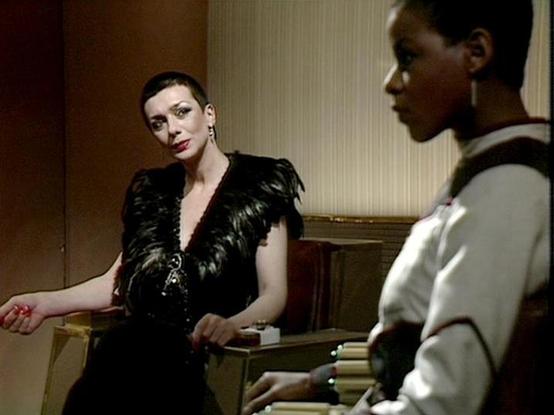 Claude Haiku 4.5 describes the image as: "# Scene Description

This image captures an indoor conversation between two women in what appears to be an elegant, formal setting. The room features warm, neutral-toned walls with classical architectural details, including what looks like decorative molding or paneling in the background.

The woman on the left wears a striking black garment with textured or feathered detailing and deep neckline, paired with statement earrings. She has dark, slicked-bac…