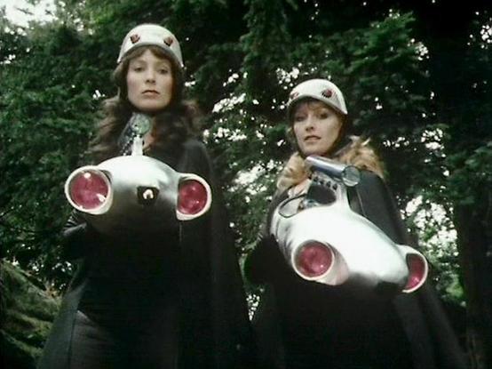 Claude Sonnet 4.5 describes the image as: "This image shows two women in a forest setting, dressed in matching white caps with badges and dark uniforms. They are posed dramatically, each holding what appears to be a futuristic white weapon or device with prominent red circular elements that resemble lights or lenses. The props have a retro-futuristic, science fiction aesthetic typical of vintage TV productions.

The scene is shot outdoors among dense foliage, with dappled sunlight filtering thr…