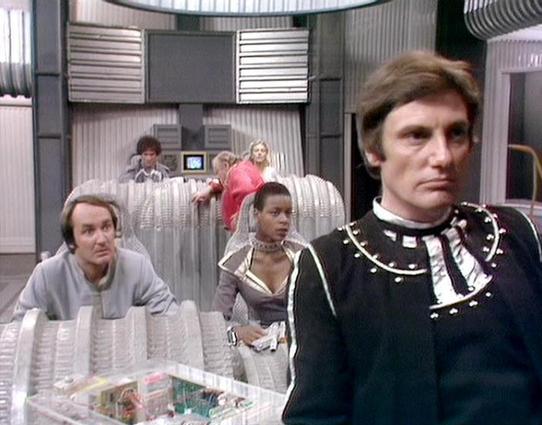 GPT 4.1 Mini describes the image as: "This image is a scene from the British science fiction television series "Blake's 7," which aired in the late 1970s and early 1980s. The setting appears to be the interior of the spaceship Scorpio, featuring a futuristic, utilitarian design with metallic and transparent elements. The characters are wearing distinctive costumes reflecting their roles within the story—ranging from military-style uniforms to more casual futuristic attire.

The actors present i…