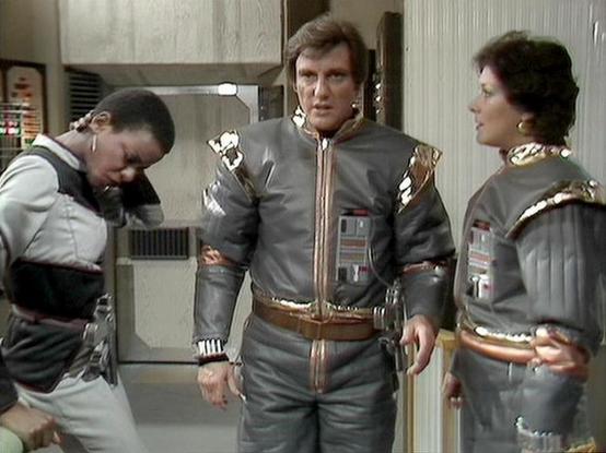 Claude Haiku 4.5 describes the image as: "# Space Station Scene

This image shows a scene set aboard a futuristic space station, featuring characters dressed in matching grey and gold uniforms typical of 1970s-80s science fiction television. The setting appears to be an interior corridor or control room with metallic walls, control panels, and utilitarian furnishings.

Three uniformed crew members are present in the scene. On the left, one character appears distressed or emotional, with their h…