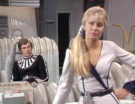 Claude Haiku 4.5 describes the image as: "# Scene Description

This image appears to be from a 1980s science fiction television production, featuring a futuristic bedroom or quarters setting. The scene shows two figures in a sleek, minimalist space with metallic furnishings and geometric design elements characteristic of the era's sci-fi aesthetic.

The woman in the foreground wears a white and black wrap-style outfit with a black belt, styled with blonde hair arranged in an elaborate braid. Sh…