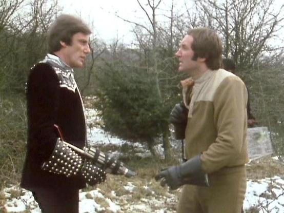 claude-opus-4-6 describes the image as: "This image shows two men in a winter outdoor setting with bare trees and patches of snow on the ground, appearing to be a scene from the British science fiction television series **Blake's 7**. 

On the left is **Paul Darrow** as **Kerr Avon**, wearing his distinctive black leather costume adorned with metal studs — a signature look for his character, the cold and calculating computer expert. On the right is **Michael Keating** as **Vila Restal**, dresse…
