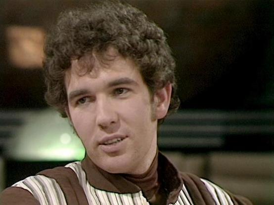 Claude Haiku 4.5 describes the image as: "# Image Description

This image shows a man with curly brown hair in what appears to be a scene from a television production. He's wearing a brown and tan striped tunic or shirt, suggesting a science fiction or period setting. The lighting and video quality indicate this is from a classic television era, likely from the 1980s or 1990s.

The setting appears to be an interior space with a blurred background, with warm lighting that creates a somewhat inti…