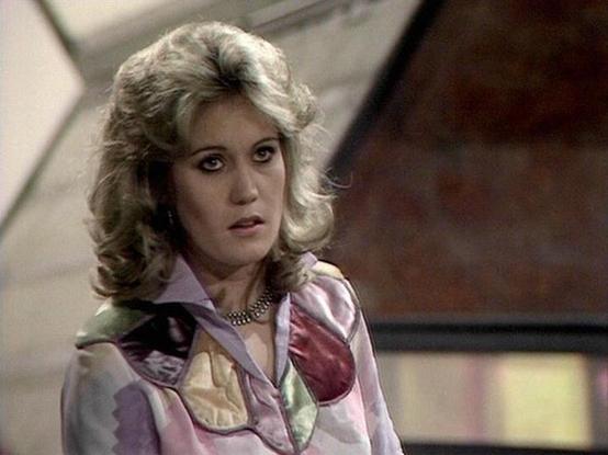 claude-opus-4-6 describes the image as: "This image shows a woman in what appears to be a science fiction television production, likely from the late 1970s or early 1980s based on the visual style, set design, and costume aesthetics. The setting features angular, futuristic-looking architecture in the background, typical of British sci-fi productions of that era.

The woman has voluminous, styled blonde hair characteristic of the period. She wears a distinctive patchwork-style blouse in soft pa…