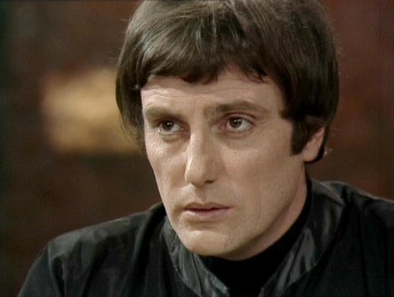 Claude Sonnet 4.5 describes the image as: "This image shows a close-up shot from what appears to be a British science fiction television production, likely from the late 1970s or early 1980s based on the film quality and styling. The subject wears a black high-necked garment and has a distinctive bowl-cut hairstyle characteristic of the era's futuristic costume design. The lighting creates a dramatic effect, emphasizing facial features against a soft, blurred background with warm brown tones.

…