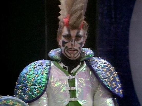 Claude Sonnet 4.5 describes the image as: "This image shows an elaborate theatrical or science fiction costume featuring striking otherworldly makeup and design. The character wears an iridescent costume with shimmering shoulder pieces in blue and green tones that catch the light dramatically. The garment appears to be a white or silver tunic with decorative fastenings down the front.

The makeup is highly stylized with bold black and white face paint creating dramatic geometric patterns across…