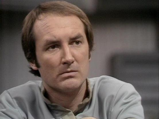 Claude Sonnet 4 describes the image as: "The image shows a man in what appears to be a **science fiction television production setting**, based on the grey, futuristic-looking costume he is wearing and the industrial/spacecraft-like background visible behind him. The set design suggests this is likely from a **late 1970s or early 1980s British sci-fi television series**, given the aesthetic style, lighting, and video quality characteristics typical of that era.

The man has **medium-length brow…