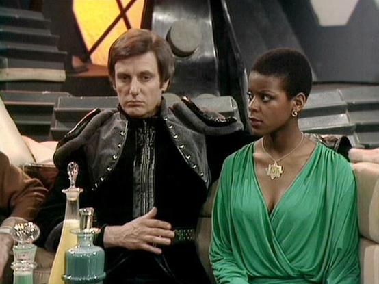 claude-opus-4-6 describes the image as: "This image shows Paul Darrow and Josette Simon in a scene from the British science fiction television series **Blake's 7**. Paul Darrow plays **Kerr Avon**, the cold, calculating anti-hero, dressed in his characteristic black leather costume with studded details and an elaborate high-collared jacket — a signature look for the character. Josette Simon plays **Dayna Mellanby**, a weapons expert who joined the crew, wearing an elegant green draped dress wit…