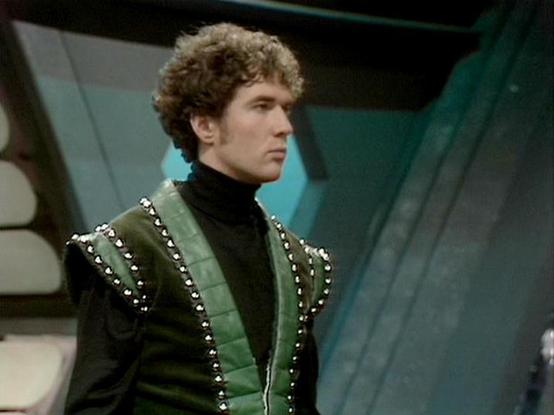 claude-opus-4-6 describes the image as: "This image shows **Steven Pacey** in his role as **Del Tarrant** from the British science fiction television series **Blake's 7**, which aired on the BBC from 1978 to 1981. Pacey joined the series in its third season as the confident and sometimes brash pilot of the Liberator spacecraft.

In the image, he is wearing one of the show's distinctive costumes — a green leather-look quilted vest with studded trim over a black turtleneck, characteristic of the …