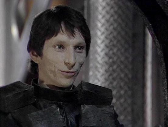 Claude Sonnet 4.5 describes the image as: "This image shows a scene from what appears to be a science fiction production, likely from the Stargate franchise based on the distinctive metallic mesh pattern visible in the background, which is characteristic of Goa'uld or Jaffa settings. The character is wearing dark, textured armor or costume pieces typical of the series' antagonistic forces. The makeup and prosthetics create a pale, otherworldly appearance with pronounced facial features. The lig…