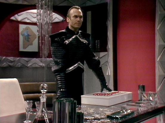 Claude Haiku 4.5 20251001 describes the image as: "# Image Description

This scene depicts a character in an elaborate black and metallic outfit standing in what appears to be a futuristic interior space. The setting features distinctive red walls and modern furnishings, including a desk with various objects and a couch visible in the background. The costume consists of a black garment with prominent metallic silver detailing and ornamental elements, suggesting a character of authority or signi…