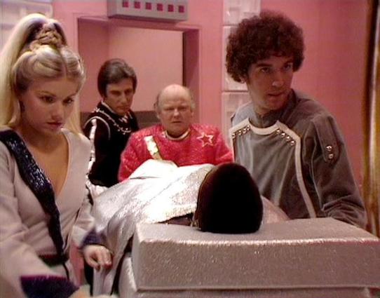 Claude Haiku 4.5 20251001 describes the image as: "# Scene Description

This image appears to be from a science fiction television production, set in a futuristic medical or laboratory environment characterized by the distinctive pink walls and clinical atmosphere. The scene shows five people gathered around what appears to be a medical examination table or pod with a white covering.

The setting suggests a space station or similar sci-fi facility, typical of 1980s television production design.…