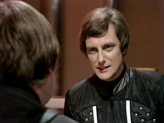 Claude Sonnet 4.5 20250929 describes the image as: "This image shows a scene from what appears to be a British science fiction television production, likely from the late 1970s or early 1980s based on the video quality and styling. The setting features a warm, brown-toned interior with two figures in conversation.

Paul Darrow is visible wearing a distinctive black leather outfit with white vertical stripes on the front, paired with a black turtleneck underneath. His hair is styled in a period-…