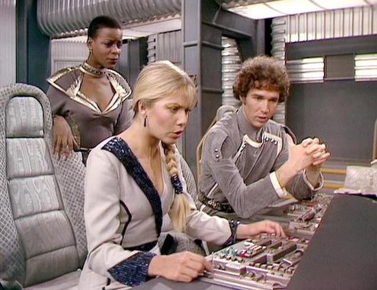 Claude Sonnet 4.5 20250929 describes the image as: "This image shows a scene from what appears to be a science fiction television production, featuring three actors in a futuristic spacecraft setting. The environment is characterized by metallic silver ducting, quilted padding on surfaces, and control panels with various buttons and switches.

In the foreground, a blonde woman in a beige uniform with black trim sits at the controls, her hair styled in a long braid. She appears focused on operat…