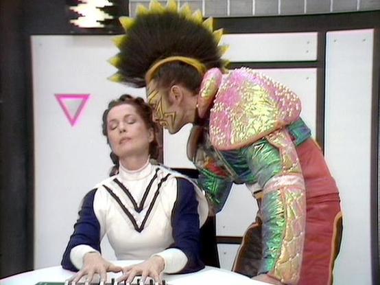 Claude Sonnet 4.5 20250929 describes the image as: "This image appears to be from a vintage science fiction television program, likely from the 1970s or 1980s. The scene shows two figures in a futuristic setting with minimalist white walls and geometric decorations, including a pink triangle symbol visible in the background.

One person is seated at what appears to be a control panel or keyboard, wearing a white and navy colored outfit with geometric V-shaped trim. The second figure stands behi…