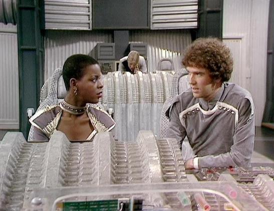 Claude Sonnet 4.6 describes the image as: "This appears to be a scene from the classic British science fiction television series **Blake's 7**, which aired on the BBC from 1978 to 1981. The image has the distinctive production style of that era, featuring futuristic set design with translucent panels, crystalline structures, and sleek grey costumes with metallic trim detailing.

The scene takes place on what appears to be the **Liberator's flight deck**, the iconic spacecraft interior central t…