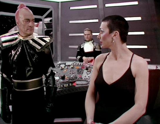 Claude Sonnet 4.6 describes the image as: "The scene appears to be from a science fiction television production, likely from the 1970s or 1980s, based on the aesthetic of the set. The setting is a spacecraft control room or bridge, featuring a futuristic console with numerous buttons and controls, along with distinctive wall panels with horizontal light strips.
Three actors are visible in the scene. On the left stands a male actor in an elaborate black villain-style costume with metallic shoul…