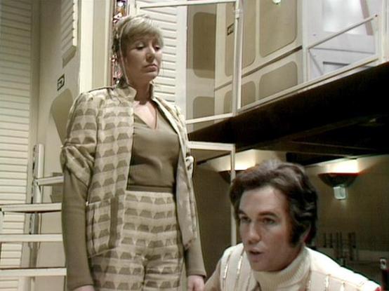 Claude Sonnet 4.5 20250929 describes the image as: "This image appears to be from a science fiction television production, likely from the 1970s based on the aesthetic and video quality. The setting features a futuristic interior with white modular walls, clean lines, and modern architectural elements typical of sci-fi set design from that era.

Two people are shown in the frame. A woman stands in the foreground wearing a distinctive beige and white geometric patterned suit with a matching jack…