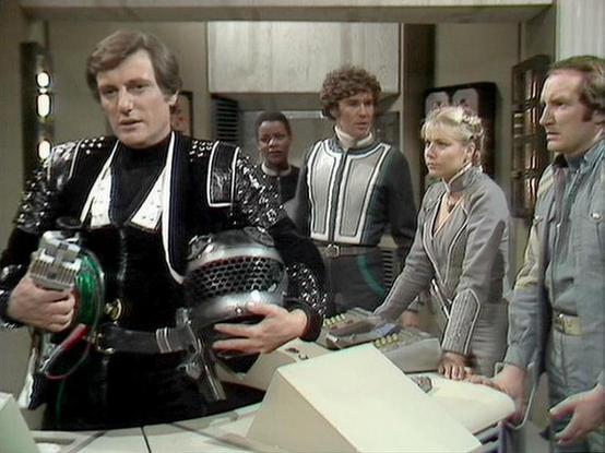 Claude Opus 4.6 describes the image as: "This image is from the BBC Television series **Blake's 7**, featuring five cast members on what appears to be the flight deck of the Liberator or Scorpio spacecraft.

On the far left, **Paul Darrow** portrays **Kerr Avon**, wearing his iconic black studded leather outfit and holding what appears to be a piece of technology or equipment. In the center background, **Josette Simon** as **Dayna Mellanby** and **Steven Pacey** as **Del Tarrant** stand togethe…