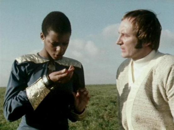 Claude Sonnet 4.6 describes the image as: "This image appears to be from the classic BBC Television series **Blake's 7**, filmed outdoors on what looks like an open moorland or grassland location under a partly cloudy sky — a setting typical of the show's location shoots.

**Josette Simon**, who played the fierce and independent warrior **Dayna Mellanby**, is seen on the left wearing her characteristic costume with distinctive gold metallic shoulder detailing and a large gold cuff bracelet. She…