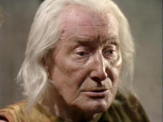 Claude Haiku 4.5 20251001 describes the image as: "# Blake's 7 Scene Description

This image captures a scene from the classic BBC Television series "Blake's 7," featuring an elderly man with long white hair and distinctive facial features. He is wearing a mustard-colored or olive-toned garment, which appears to be period costume appropriate to the science fiction setting of the series.

The actor displays a contemplative or serious expression, characteristic of dramatic moments in the show. Th…