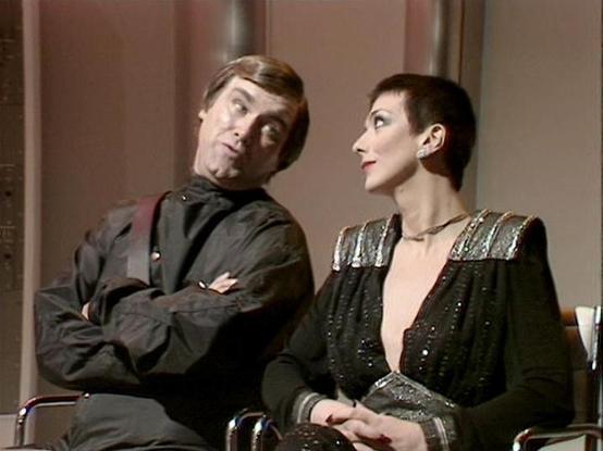 Claude Haiku 4.5 20251001 describes the image as: "# Blake's 7 Scene

This scene from the classic BBC Television series "Blake's 7" depicts two characters in what appears to be an interior spacecraft setting, characterized by the muted earth-tone walls typical of the show's production design. The male character on the left wears a dark military-style jacket with a distinctive emblem, suggesting a position of authority within the Federation hierarchy. His posture is relaxed yet commanding, with …