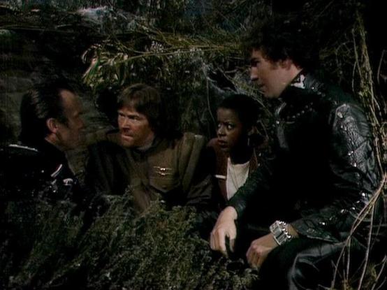 Claude Haiku 4.5 20251001 describes the image as: "# Blake's 7 Scene Description

This image captures a tense moment from the classic BBC Television series "Blake's 7," showing a group of characters huddled together in what appears to be an underground or enclosed setting. The scene is dimly lit, with overgrown vegetation and moss-covered surfaces visible around them, suggesting they may be hiding or taking shelter in a concealed location.

The five individuals are dressed in the distinctive fa…