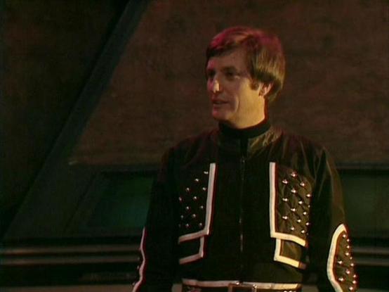 Claude Opus 4.6 describes the image as: "This image shows Paul Darrow in his iconic role as Kerr Avon from the classic BBC Television series "Blake's 7." He is wearing one of Avon's distinctive costumes — a black leather-look jacket featuring studded detailing, white/cream trim accents, and a front zipper, which is characteristic of the show's bold, futuristic costume design. The outfit reflects Avon's sharp, stylish persona that set him apart from other characters in the series.

Darrow is cap…