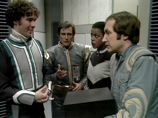 Claude Opus 4.6 describes the image as: "This image is from the BBC Television series *Blake's 7*. The scene shows four crew members of the Liberator (later Scorpio) engaged in what appears to be an intense discussion or debate aboard their spacecraft.

On the far left stands Steven Pacey as Del Tarrant, wearing a distinctive green and white tunic, his curly hair characteristic of the role. He appears to be making a point to the group. In the center is Paul Darrow as Kerr Avon, dressed in his s…