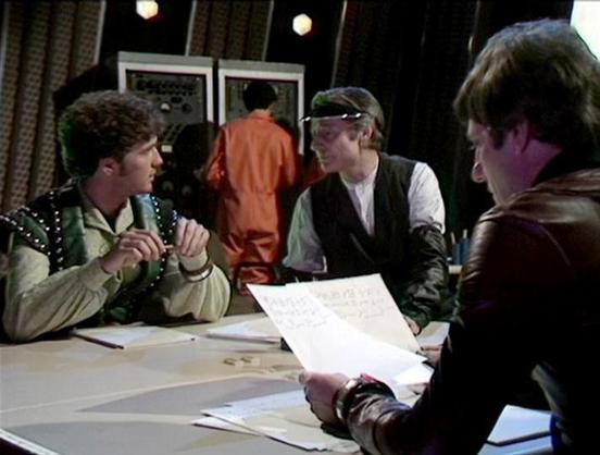 Claude Haiku 4.5 20251001 describes the image as: "# Blake's 7 Scene

This scene depicts an intense meeting in what appears to be a spacecraft interior or command center. The setting features a modern, minimalist design typical of the science fiction aesthetic of the series, with white panels, curtains, and overhead lighting creating a clinical atmosphere.

Several characters are gathered around a table with documents and papers spread before them, suggesting a strategic briefing or planning se…