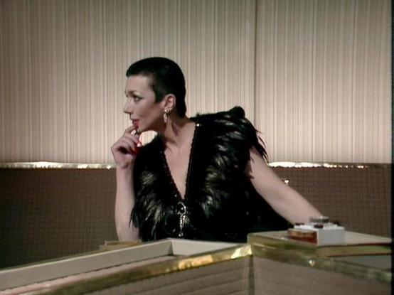 Claude Sonnet 4.5 20250929 describes the image as: "This image captures a dramatic scene from the classic BBC Television series "Blake's 7." The setting appears to be an elegant, modernist interior with distinctive striped wall paneling featuring vertical lines in cream and beige tones. A horizontal strip of lighting creates an atmospheric glow across the middle of the wall.

Jacqueline Pearce commands the frame in this scene, positioned at what appears to be a sleek desk or console. She wears …