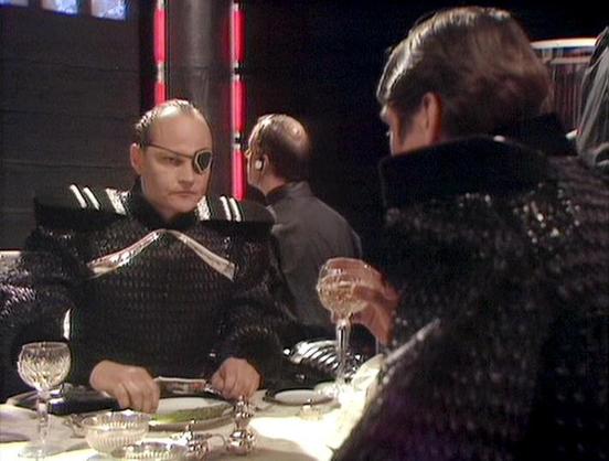 Claude Opus 4.6 describes the image as: "This image is from the classic BBC Television series **Blake's 7**. The scene depicts a formal dining setting with a dark, atmospheric aesthetic typical of the show's production design. 

On the left sits **David Quilter**, wearing a distinctive black and silver costume with an eye patch, giving his character a menacing, authoritarian appearance. His costume features metallic accents and structured shoulder details characteristic of the show's futuristic…