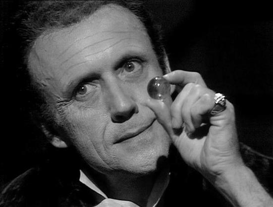 Claude Haiku 4.5 20251001 describes the image as: "# Black and White Portrait from Blake's 7

This is a black and white promotional or scene photograph from the classic BBC Television series "Blake's 7." The image captures a man in formal attire, wearing a dark jacket and white shirt, photographed against a stark black background. He displays a characteristic expression—a slight, knowing smile with a raised eyebrow—while holding what appears to be a small spherical object near his face, examini…