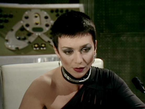 Claude Sonnet 4.5 20250929 describes the image as: "This image shows a scene from the classic BBC Television series "Blake's 7". The photograph captures an elegant figure seated in a white chair, wearing a striking black one-shoulder garment with metallic silver detailing around the neckline. The styling is distinctly bold and theatrical, featuring a short dark pixie haircut, dramatic eye makeup, and deep red lipstick that emphasizes the character's commanding presence.

The setting appears to …