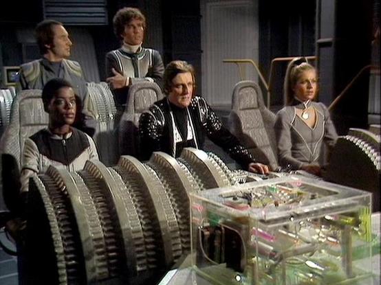 Claude Sonnet 4.6 describes the image as: "This image is from the classic BBC Television series **Blake's 7**, likely from a later series given the distinctive Scorpio spacecraft interior setting. The scene takes place on the flight deck, recognizable by its characteristic ribbed/ridged seating arrangement and the transparent equipment console visible in the foreground.

The five crew members are seated in the spacecraft's distinctive molded crew stations. The set design reflects the show's sig…
