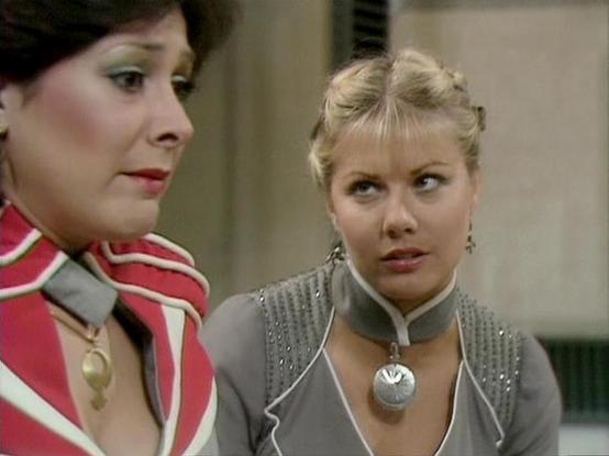 Claude Haiku 4.5 20251001 describes the image as: "# Blake's 7 Scene

This scene from the classic BBC Television series "Blake's 7" depicts two women in what appears to be an interior spacecraft or station setting. The woman on the left wears a striking red and white striped military-style uniform with a gold pendant, her dark hair styled in a short, sleek cut. She gazes thoughtfully to the side, her expression contemplative.

The woman on the right has blonde hair styled in an upswept fashion …