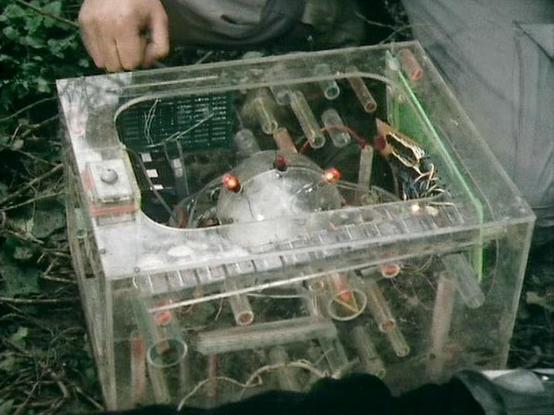 Claude Haiku 4.5 20251001 describes the image as: "# Blake's 7 - Technical Equipment Scene

This image shows a transparent acrylic box containing various electronic components and circuitry, photographed outdoors on grass. The device appears to be some form of specialized equipment being examined or operated by crew members visible in the frame.

The transparent casing allows viewers to see the internal mechanisms, including circuit boards, wiring, metallic components, and what appear to be ind…