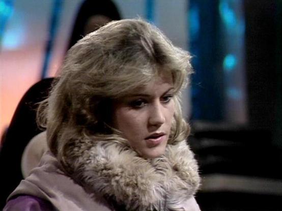 Claude Sonnet 4.6 describes the image as: "This image appears to be a still from the classic BBC Television series **Blake's 7**, likely from the late 1970s or early 1980s, based on the production style and aesthetic.

The setting has the characteristic look of a science fiction TV production of that era, with colorful studio lighting visible in the background — including blue and orange tones — suggesting an interior spacecraft or futuristic environment set.

The actress in the image is wearin…