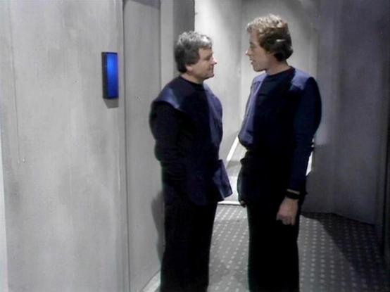 Claude Haiku 4.5 20251001 describes the image as: "# Blake's 7 Scene

This image captures a tense moment in a stark, minimalist corridor aboard a spacecraft. Two men in dark uniforms stand face-to-face in what appears to be an interior ship passage, characterized by plain grey walls and a simple floor with a textured pattern. A small blue illuminated panel is visible on the wall to the left, suggesting advanced technology typical of the series' aesthetic.

The setting reflects the utilitarian d…