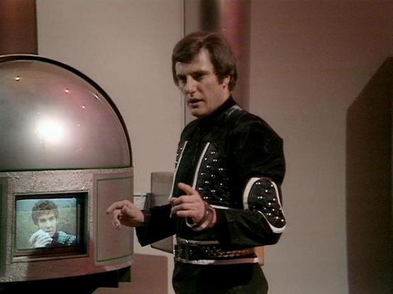 Claude Sonnet 4.5 20250929 describes the image as: "This image shows a scene from the classic BBC Television series "Blake's 7". The actor Paul Darrow is depicted wearing a distinctive black leather costume with metallic studs and white trim details characteristic of the show's futuristic aesthetic. He is holding what appears to be a cup while standing next to a retro-futuristic communication or viewing device with a domed top. The device features a small screen displaying another person's face…