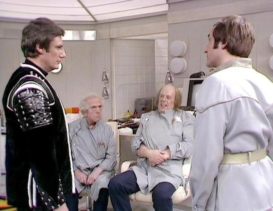 Claude Sonnet 4.5 20250929 describes the image as: "This image shows a scene from the classic BBC Television series "Blake's 7" set in what appears to be a medical bay or laboratory. The sterile, white-walled environment features clinical furnishings and equipment typical of the show's spacecraft interiors.

Four actors are present in the scene. On the left stands Paul Darrow wearing the distinctive black leather costume with silver studs that became iconic for his character. To the right is Mi…
