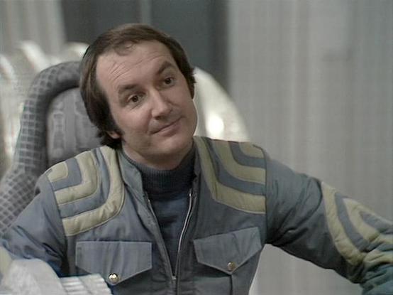 Claude Sonnet 4.5 20250929 describes the image as: "This image shows a scene from the classic BBC Television series "Blake's 7". The actor Michael Keating is seated in what appears to be a spacecraft interior, characterized by the grey padded seating visible behind him. He's wearing a distinctive grey and cream colored quilted vest or jacket with horizontal striping over a dark turtleneck sweater. His expression is somewhat quizzical or bemused, with raised eyebrows and a slight smile.

The set…