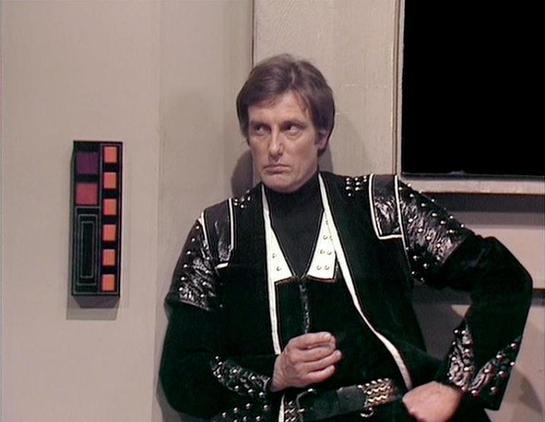 Claude Haiku 4.5 20251001 describes the image as: "# Blake's 7 Scene Description

This image captures a scene from the classic BBC Television series "Blake's 7," featuring an actor in an elaborate costume characteristic of the show's science fiction aesthetic. The character wears a striking black velvet jacket adorned with ornate silver embroidery and studded details across the shoulders and sleeves. A distinctive white collar frames the neckline, and a dark chain necklace completes the ensembl…