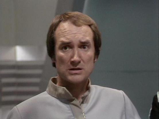 Claude Sonnet 4.5 20250929 describes the image as: "This image shows Michael Keating in character from the BBC television series "Blake's 7". He appears in a beige or tan colored tunic with a characteristic mandarin-style collar, which was typical of the show's futuristic costume design. The shot is a close-up capturing his facial expression, which conveys concern or apprehension. His brown hair is styled in the fashion typical of the late 1970s when the series was produced.

The setting appear…