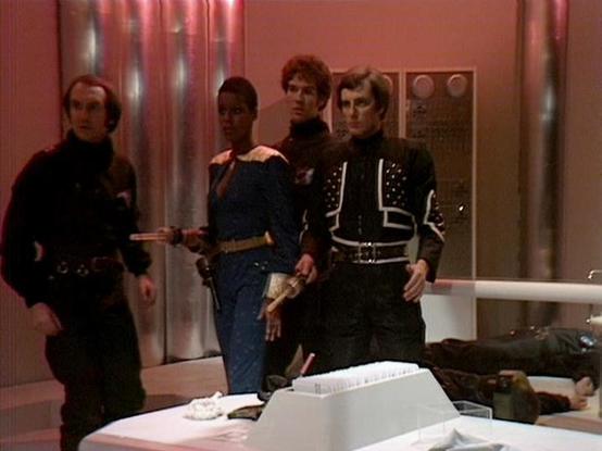 Claude Sonnet 4.6 describes the image as: "This image is from the classic BBC Television series **Blake's 7**, depicting a tense scene set aboard what appears to be a spacecraft interior. The set features the characteristic design of the era — metallic silver wall panels, reddish ambient lighting, and futuristic control surfaces in the foreground.

Four characters are visible standing together in a confrontational or cautious grouping. One character wears a distinctive **black jacket with white…