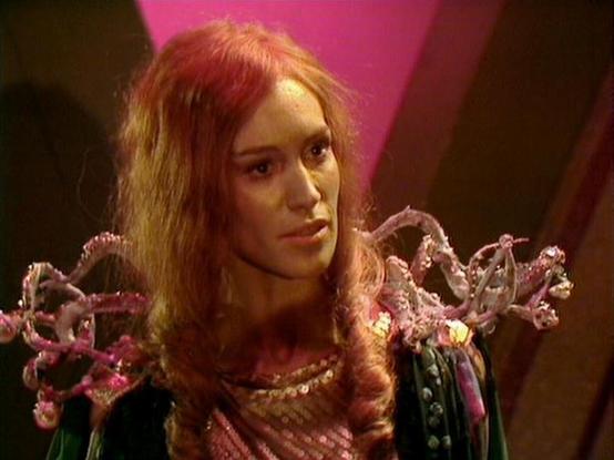 Claude Sonnet 4.6 describes the image as: "This image is from the classic BBC Television series **Blake's 7**, which aired from 1978 to 1981. The scene appears to be set aboard a spacecraft or alien environment, characteristic of the show's science fiction aesthetic.

The setting features the distinctive low-budget but imaginative production design typical of the era, with a pink/magenta background and dramatic lighting. The costume design is particularly notable — the character is wearing an e…