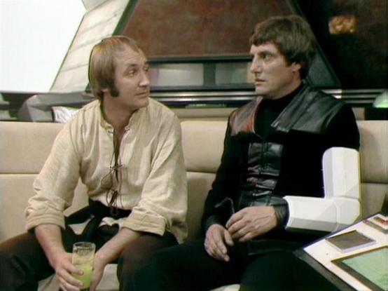 Claude Sonnet 4.5 20250929 describes the image as: "This image shows two characters in a futuristic spacecraft setting from the classic BBC Television series "Blake's 7". The scene takes place in what appears to be a relaxation or briefing area, with cream-colored seating and modern, sleek interior design typical of the show's aesthetic.

On the left, Michael Keating wears a casual cream-colored tunic with a black sash or belt, holding what appears to be a glass with a yellow beverage. His char…