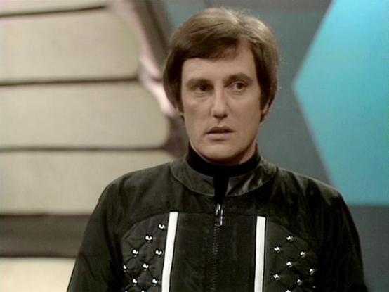 Claude Haiku 4.5 20251001 describes the image as: "# Blake's 7 - Studio Scene

This image captures a moment from the classic BBC science fiction series "Blake's 7," showing an actor in character wearing distinctive costume elements typical of the show's production design. The setting appears to be a studio set with a futuristic aesthetic, featuring geometric shapes and a turquoise background panel that exemplifies the 1970s sci-fi television visual style.

The character is dressed in a black ja…